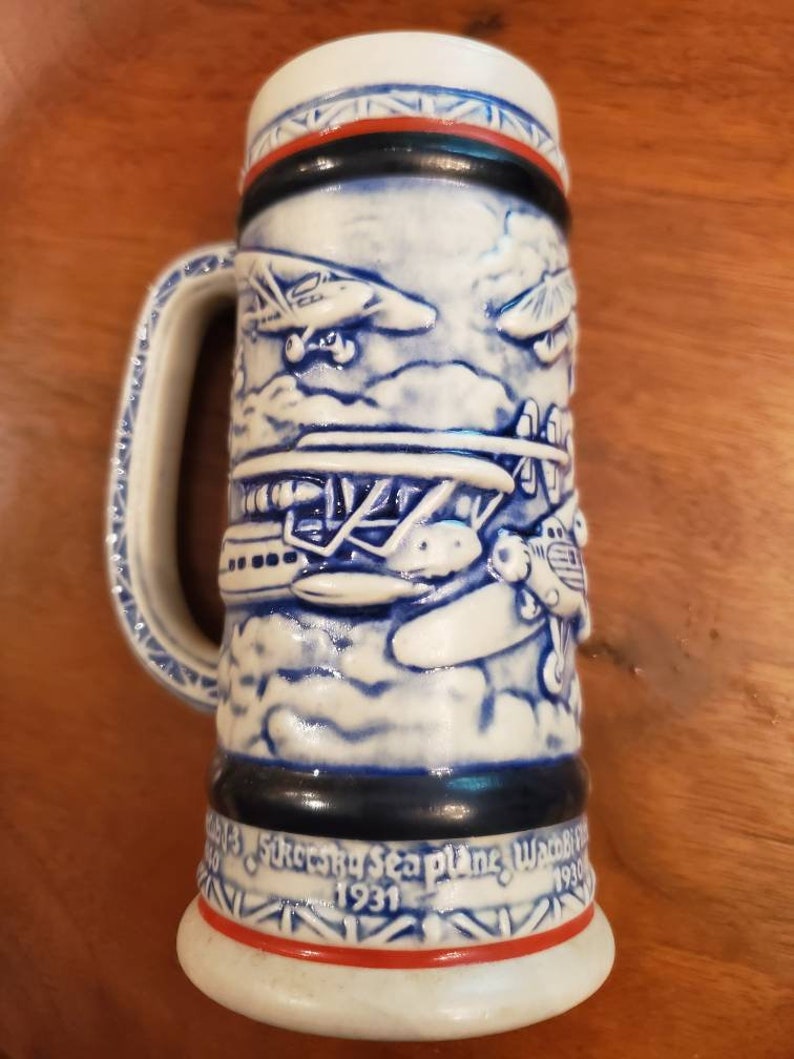 Pode incluir: Uma caneca de cerveja de cer&acirc;mica branca com detalhes em azul e vermelho. A caneca apresenta um design em relevo de hidroavi&otilde;es antigos voando nas nuvens. O texto "Sikorsky Seaplane Waterbury 1931" est&aacute; impresso na parte inferior da caneca.
