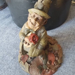 May include: A whimsical figurine of a smiling man in a green coat and hat, holding a red rose. The figure is seated on a base with leaves and acorns. The figurine is approximately 4 inches tall.