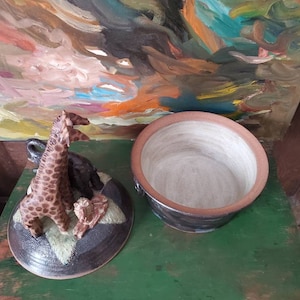 May include: A ceramic lidded dish with a brown glaze and a scene of a giraffe, a lion, and an elephant sculpted on the lid. The dish is empty and has a white interior.