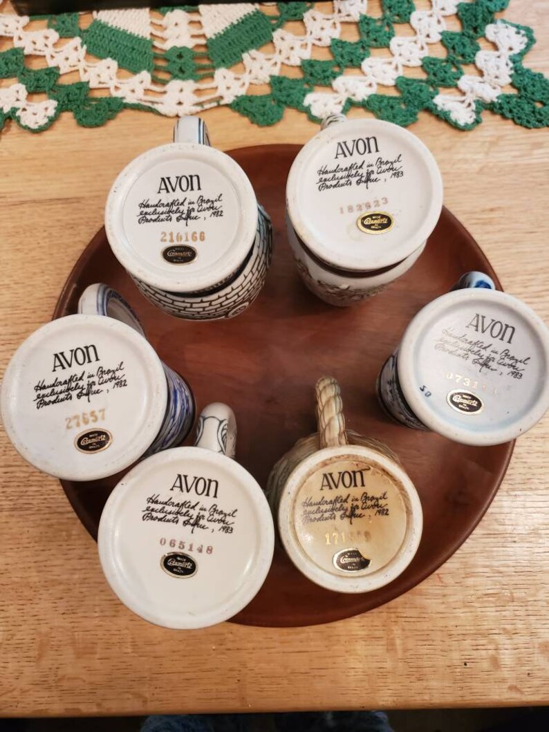 Pode incluir: Seis canecas de cer&acirc;mica branca com detalhes azuis e dourados. Cada caneca tem um design diferente e apresenta um r&oacute;tulo manuscrito na parte inferior que diz "AVON Handcrafted in Germany exclusively for Avon Products Office, 1983" e um n&uacute;mero &uacute;nico.