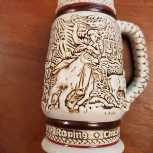 Pode incluir: Uma caneca de cerveja de cer&acirc;mica branca com uma al&ccedil;a de corda marrom. A caneca apresenta um design em relevo detalhado de um cowboy la&ccedil;ando um touro. A parte inferior da caneca diz "Roping Chuck"