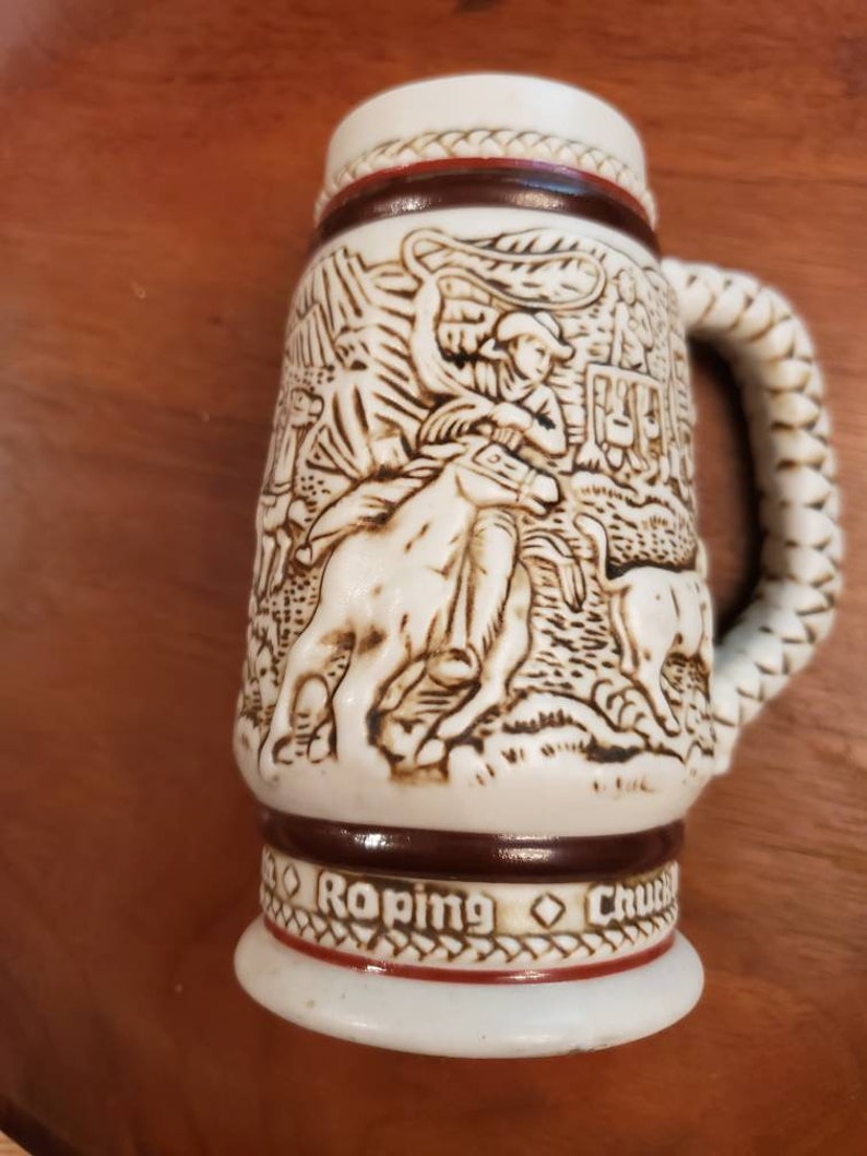 Pode incluir: Uma caneca de cerveja de cer&acirc;mica branca com uma al&ccedil;a de corda marrom. A caneca apresenta um design em relevo detalhado de um cowboy la&ccedil;ando um touro. A parte inferior da caneca diz "Roping Chuck"