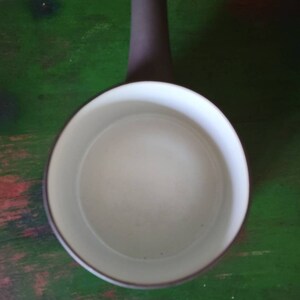 Op de afbeelding: Een witte keramische kan met een grijze handgreep. De kan heeft een rond lichaam en een brede opening.