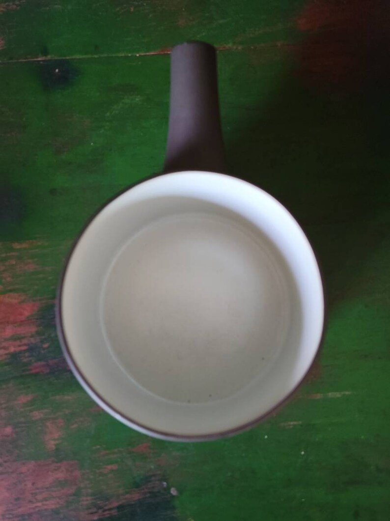 Op de afbeelding: Een witte keramische kan met een grijze handgreep. De kan heeft een rond lichaam en een brede opening.