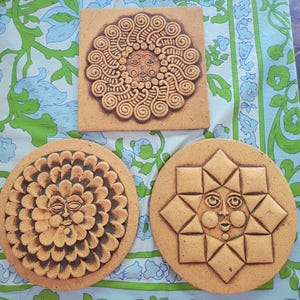 May include: Three ceramic tiles with sun-like faces. One is square with a swirling design, and two are round with geometric and floral patterns. The tiles are tan with brown detailing, set against a patterned blue and green fabric.