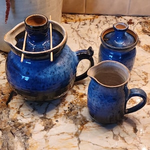 May include: A set of three blue and white ceramic kitchenware items. A teapot with a wooden lid and a wooden handle, a small jar with a lid, and a pitcher with a handle.