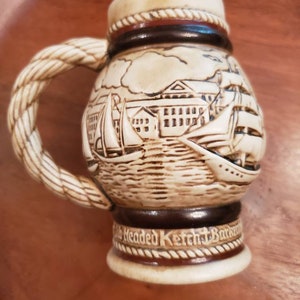 Pode incluir: Uma caneca de cerveja de cer&acirc;mica com uma al&ccedil;a em forma de corda e uma cena detalhada de um porto com navios e edif&iacute;cios. A caneca &eacute; decorada com detalhes marrons e brancos e tem o texto "Old Headed Ketch Barkentine" na parte inferior.