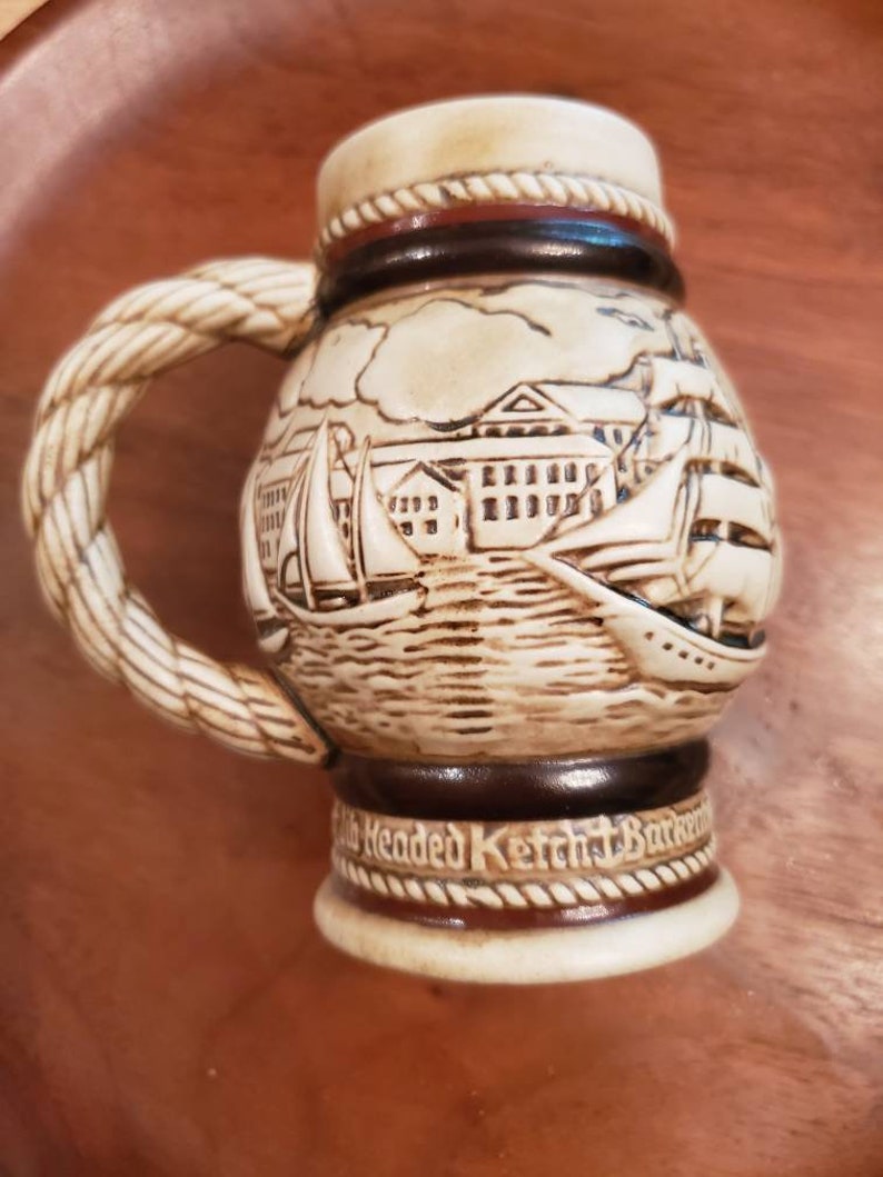 Pode incluir: Uma caneca de cerveja de cer&acirc;mica com uma al&ccedil;a em forma de corda e uma cena detalhada de um porto com navios e edif&iacute;cios. A caneca &eacute; decorada com detalhes marrons e brancos e tem o texto "Old Headed Ketch Barkentine" na parte inferior.