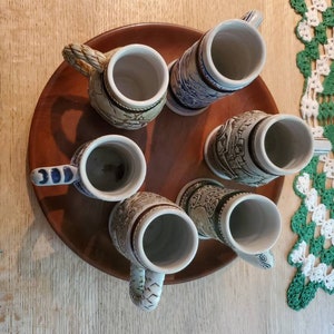 Pode incluir: Um conjunto de seis canecas de cerveja de cer&acirc;mica com v&aacute;rios desenhos, incluindo um castelo, um navio e uma cena de montanha. As canecas est&atilde;o dispostas em uma bandeja de madeira.