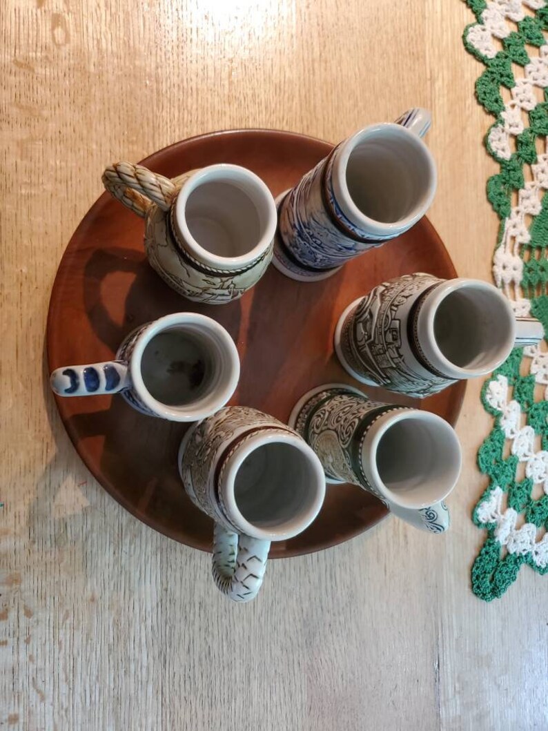Pode incluir: Um conjunto de seis canecas de cerveja de cer&acirc;mica com v&aacute;rios desenhos, incluindo um castelo, um navio e uma cena de montanha. As canecas est&atilde;o dispostas em uma bandeja de madeira.