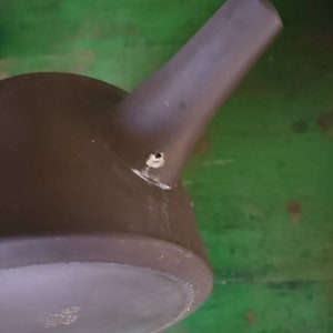 Op de afbeelding: Een donkergrijze theepot van keramiek met een tuit en een klein gaatje aan de zijkant. De theepot staat op een groene ondergrond.