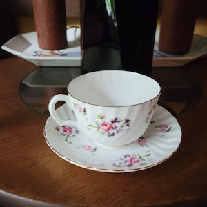 May include: A white teacup and saucer with a floral pattern in shades of pink, purple, and green. The saucer has a gold trim.