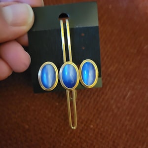 May include: A gold-toned hair clip and matching stud earrings set. Each piece features an oval, blue cabochon stone with a gold-colored border. The set is displayed on a black card.