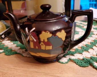 May include: A brown teapot with a windmill and houses painted on it. The teapot has a lid and a handle.