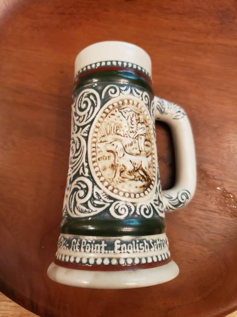 Pode incluir: Uma caneca de cerveja de cer&acirc;mica com um design verde e branco. A caneca apresenta uma imagem em relevo de um c&atilde;o em uma cena de floresta. A parte inferior da caneca diz "St. Point... English Setter".