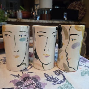 May include: Three cylindrical ceramic tumblers with an off-white base color. Each tumbler features hand-painted abstract facial features in black lines, accented with soft patches of yellow, blue, purple, and green paint across the surfaces.