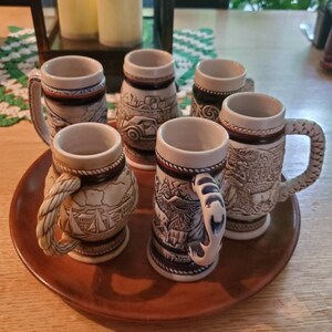 Pode incluir: Conjunto de seis canecas de cerveja de cer&acirc;mica com v&aacute;rios desenhos, incluindo um veleiro, uma cena de montanha e um carro. As canecas s&atilde;o brancas com detalhes azuis e marrons e t&ecirc;m uma al&ccedil;a em forma de corda.