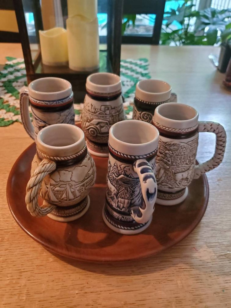 Pode incluir: Conjunto de seis canecas de cerveja de cer&acirc;mica com v&aacute;rios desenhos, incluindo um veleiro, uma cena de montanha e um carro. As canecas s&atilde;o brancas com detalhes azuis e marrons e t&ecirc;m uma al&ccedil;a em forma de corda.