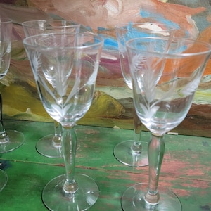 May include: Set of six clear glass wine glasses with etched wheat designs. The glasses have a tall, slender stem and a round bowl. The glasses are arranged on a green wooden surface.