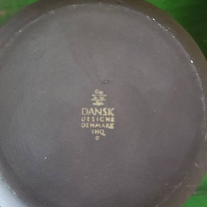 Op de afbeelding: De onderkant van een donkergrijze keramische theepot met een gouden inscriptie die "DANSK DESIGN DENMARK IMQ" luidt.
