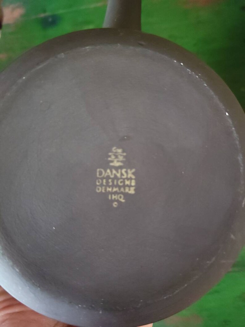 Op de afbeelding: De onderkant van een donkergrijze keramische theepot met een gouden inscriptie die "DANSK DESIGN DENMARK IMQ" luidt.