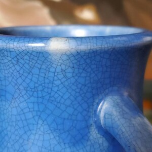 May include: Close-up of a blue ceramic vase with a crackled glaze. The vase has a white rim and a white handle.