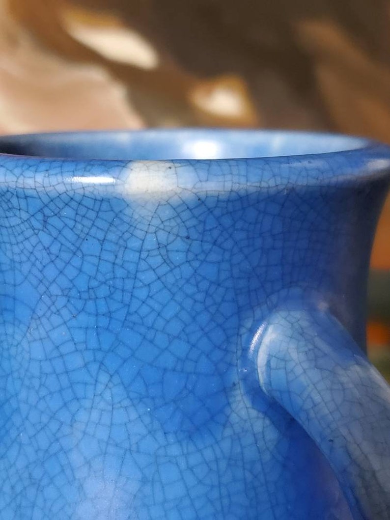 May include: Close-up of a blue ceramic vase with a crackled glaze. The vase has a white rim and a white handle.