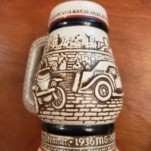 Pode incluir: Uma caneca de cerveja de cer&acirc;mica branca com um design marrom e preto de um carro antigo. A caneca tem uma faixa azul e vermelha ao redor da parte superior e o texto "Day Streamer - 1936 MG - 197" na parte inferior.