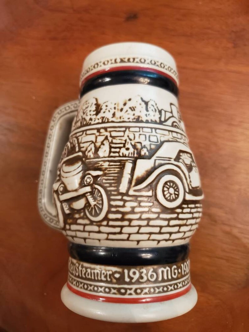 Pode incluir: Uma caneca de cerveja de cer&acirc;mica branca com um design marrom e preto de um carro antigo. A caneca tem uma faixa azul e vermelha ao redor da parte superior e o texto "Day Streamer - 1936 MG - 197" na parte inferior.