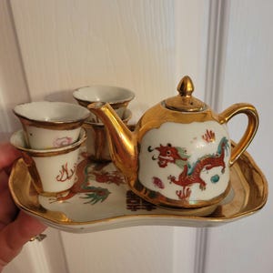 May include: A gold and white porcelain tea set with a dragon design. The set includes a teapot, four cups, and a tray.