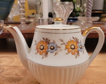 Op de afbeelding: Een witte theepot met een gouden rand en een bloemenpatroon. De theepot heeft een deksel met een gouden knop en een tuit aan de zijkant. De theepot staat op een houten oppervlak.
