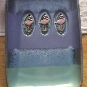 May include: A rectangular ceramic tray with a blue and green glaze. The tray features three raised oval designs with pink flamingos.