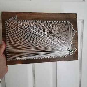 May include: A wooden string art piece depicting the outline of Pennsylvania. White string is stretched across the wooden board, creating a geometric design. The wood has a dark brown stain and is rectangular.