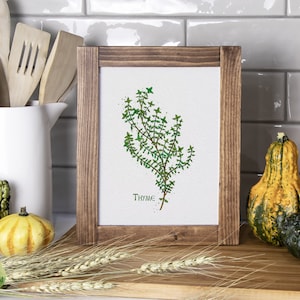 Puede incluir: Una pintura acuarela enmarcada de una rama de tomillo con la palabra "THYME" escrita debajo. El marco es de madera marrón y la pintura tiene un fondo blanco. La pintura está sobre una superficie de madera con calabazas y tallos de trigo.