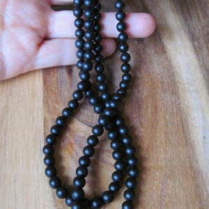 May include: A black beaded necklace with a matte finish. The necklace is composed of numerous small, round beads strung together. It features a silver-colored clasp. The necklace is displayed against a wooden background.