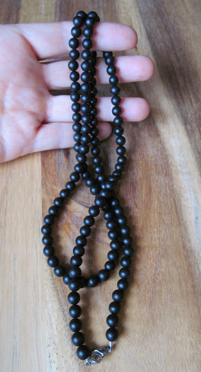May include: A black beaded necklace with a matte finish. The necklace is composed of numerous small, round beads strung together. It features a silver-colored clasp. The necklace is displayed against a wooden background.