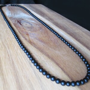 May include: A black beaded necklace with a matte finish. The necklace is composed of small, round beads and features a silver-colored clasp. The beads are evenly spaced, creating a simple and elegant design. The necklace is displayed on a wooden surface.