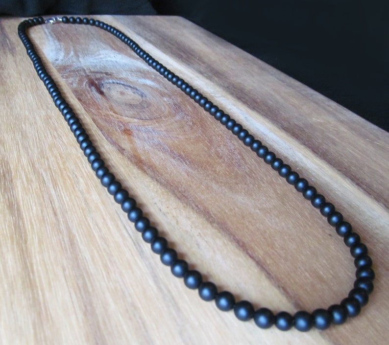 May include: A black beaded necklace with a matte finish. The necklace is composed of small, round beads and features a silver-colored clasp. The beads are evenly spaced, creating a simple and elegant design. The necklace is displayed on a wooden surface.