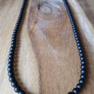 May include: A black beaded necklace with a matte finish. The beads graduate in size, with larger beads at the front and smaller ones towards the back. The necklace is displayed on a wooden surface.