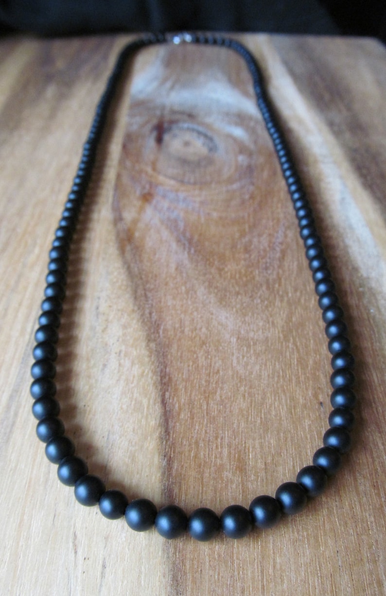 May include: A black beaded necklace with a matte finish. The beads graduate in size, with larger beads at the front and smaller ones towards the back. The necklace is displayed on a wooden surface.