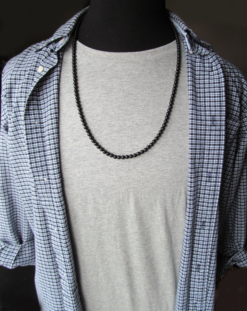 May include: A black beaded necklace draped over a gray t-shirt. The necklace is composed of small, round beads. The t-shirt is worn under an open, blue and white plaid button-down shirt.