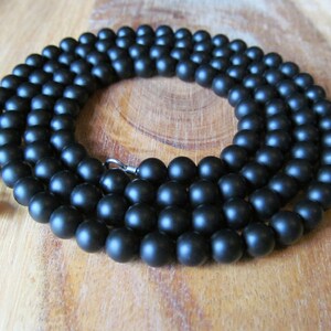 May include: A matte black beaded necklace. The necklace is composed of numerous round, dark beads strung together. A silver-colored clasp secures the necklace. The beads are approximately 8mm in diameter.