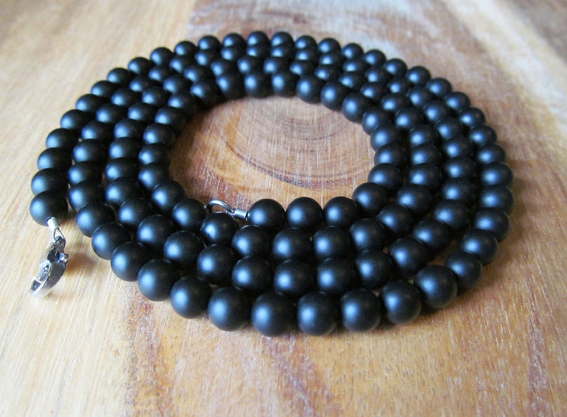 May include: A matte black beaded necklace. The necklace is composed of numerous round, dark beads strung together. A silver-colored clasp secures the necklace. The beads are approximately 8mm in diameter.