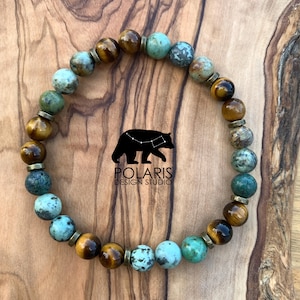 Puede incluir: Una pulsera de cuentas con una mezcla de cuentas turquesas, marrones y doradas. La pulsera está hecha de cuentas de piedra natural y está ensartada en un cordón elástico. El logotipo de Polaris Design Studio es visible en el fondo.