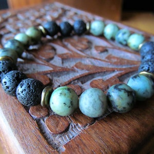 May include: A beaded bracelet featuring a mix of natural stone beads. The bracelet includes dark gray lava rock beads, light green and blue-green stone beads, and gold-colored spacers. The bracelet is displayed on a carved wooden surface.