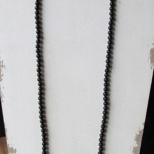 May include: A long, black beaded necklace displayed on a white, distressed wooden stand. The necklace is composed of small, round beads, creating a simple yet elegant accessory. The beads appear to be matte.