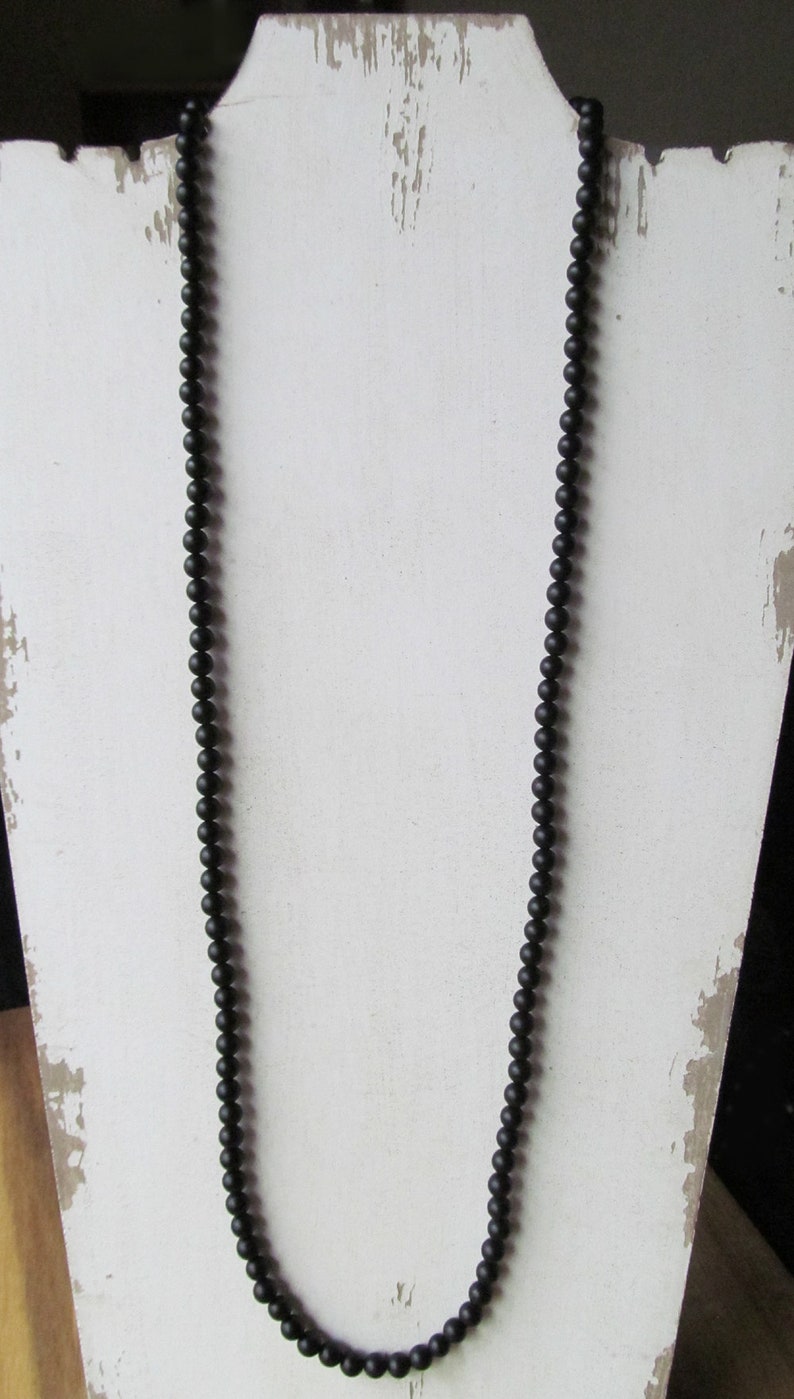 May include: A long, black beaded necklace displayed on a white, distressed wooden stand. The necklace is composed of small, round beads, creating a simple yet elegant accessory. The beads appear to be matte.