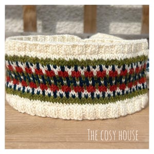 May include: Cream-colored knitted headband with a geometric pattern in red, green, and navy blue. The headband is made of a thick, textured yarn and is designed to keep the ears warm. The text "THE COSY HOUSE" is at the bottom.