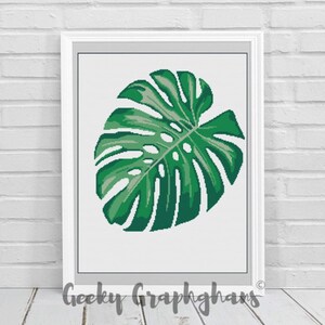 Könnte beinhalten: Ein gerahmtes Kreuzstichmuster mit einem einzelnen grünen Monstera-Blatt auf weißem Hintergrund. Das Muster trägt den Titel "Geeky Graphghans".