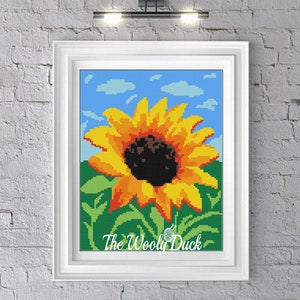 Puede incluir: Un patrón de punto de cruz con un girasol amarillo brillante con un centro marrón oscuro, sobre un fondo de cielo azul con nubes blancas. El girasol está rodeado de hojas y tallos verdes. El patrón se titula "The Wooly Duck".